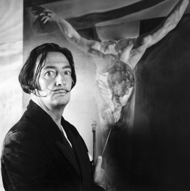 Featured Image: Spanish Surrealist painter Salvador Dalí in his studio in Port Lligat with his painting of Christ on the cross. salvador dali exhumed
