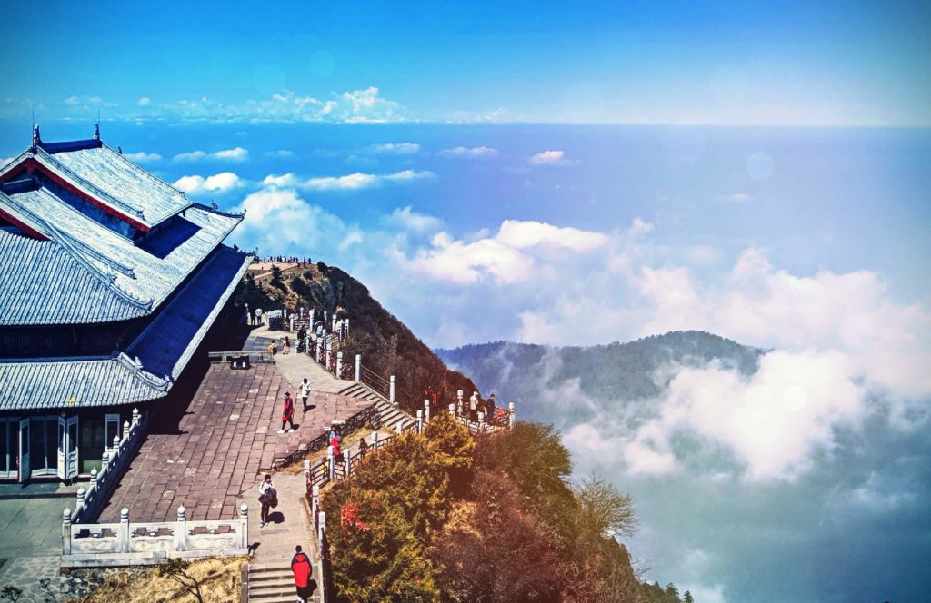 the Buddhist temple is perching on the edge of the mountain, people are walking up the staircase and on the terrace in the foreground, cloudy sky is in the distance