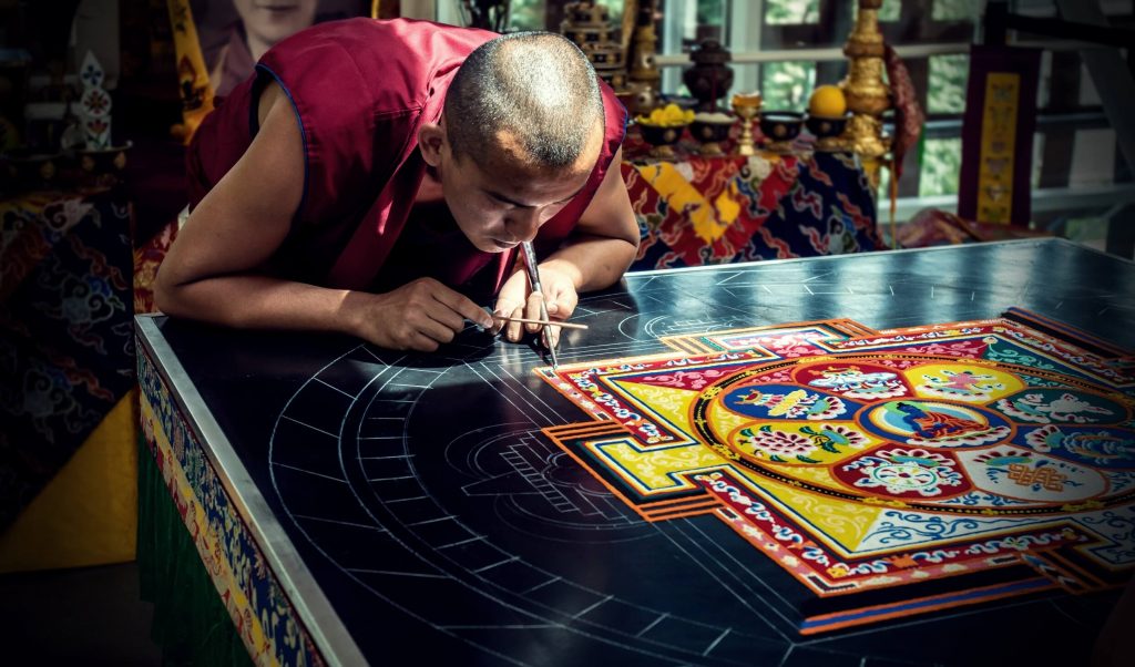 the Buddhist monk is leaning and constructs the colorful sand mandala on the table