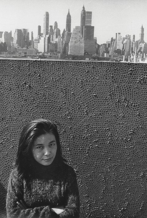 Black and white photograph of Yayoi Kusama taken from above. A large painting taking all the picture stands behind her, while New York skyline is in the background.
