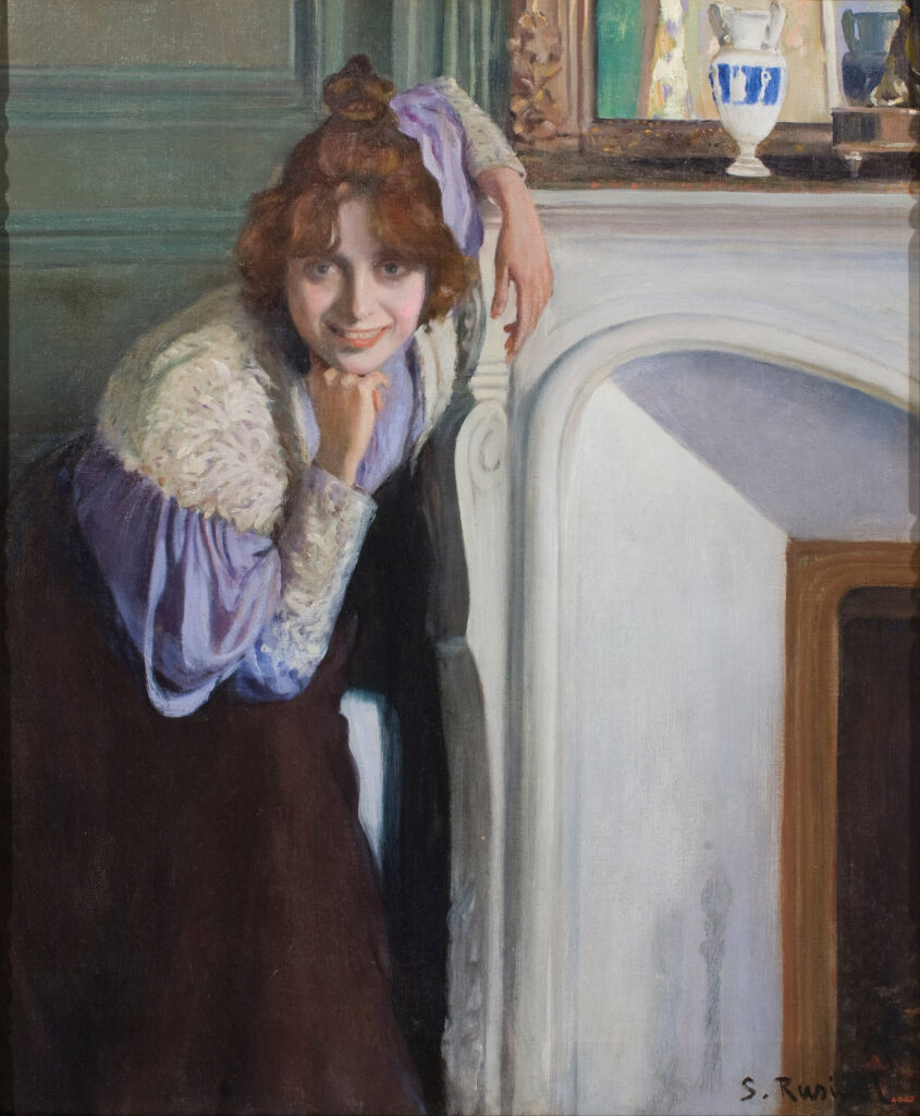 A white woman with dark blonde hair stares at the viewer; one elbow leans on a fireplace mantle; her chin rests on her elbow which rests on her knee.