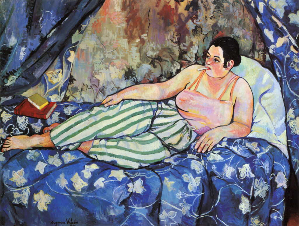 Suzanne Valadon Barnes Foundation: A white woman lays on a bed wearing a pink camisole and green and white striped pants. She has an unlit cigarette in her mouth and books at her feet. The bed curtains behind her are open and have a blue and white pattern, as does the bed spread.