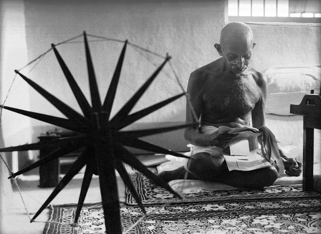 women photographers, Margaret Bourke-White, Mohandas Gandhi reads as he sits cross-legged on the floor next to a spinning wheel at home, 1946.