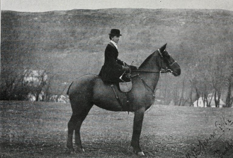 Black and white photograph of a caucasian woman astride a horse, facing right