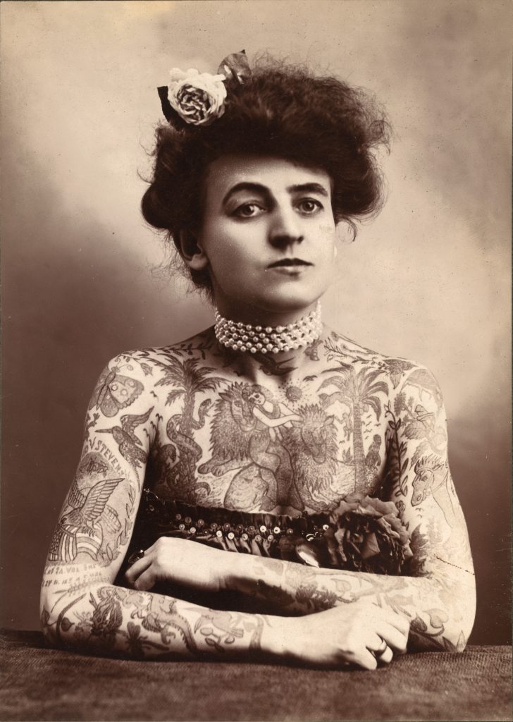 A black and white photograph of a Caucasian woman leaning on a table with her arms crossed in front of her. She's confident and badass looking at the camera. She's wearing a bodice that reveals her chest and arm tattoos.