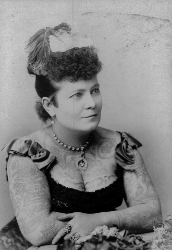 A black and white photograph of a Caucasian woman leaning forward on a table. She stares to the side of camera. She's wearing a bodice and her arms, chest, and neck is tattooed.