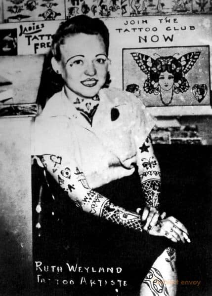 A black and white photograph of a Caucasian woman sitting cross legged in a tattoo parlor holding a cigarette and looking at the camera. She wears a white blouse and black skirt. She has tattoos on her arms, legs, and face.