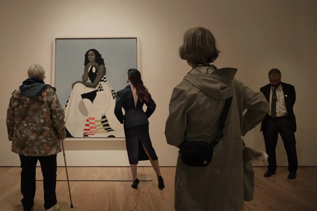 Anne hathaway photoshoot: Anne Hathaway looking at the Amy Sherald’s Michelle LaVaughn Robinson Obama at the Whitney Museum of American Art. Photograph by Annie Leibovitz. Vogue.
